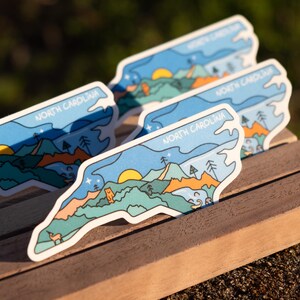 May include: Several stickers with a North Carolina design. Each sticker features a landscape with mountains, trees, a sun, and the text "NORTH CAROLINA" in a blue and white color scheme. The stickers are on a wooden surface.
