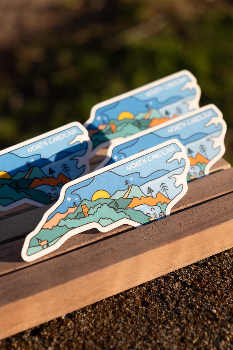 May include: Several stickers with a North Carolina design. Each sticker features a landscape with mountains, trees, a sun, and the text "NORTH CAROLINA" in a blue and white color scheme. The stickers are on a wooden surface.