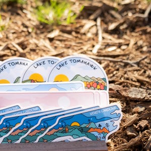 May include: A stack of stickers featuring landscape designs with text. The stickers have a variety of shapes and colors, including blue, pink, and white. Some stickers say "Lake Tomahawk" and "North Carolina".