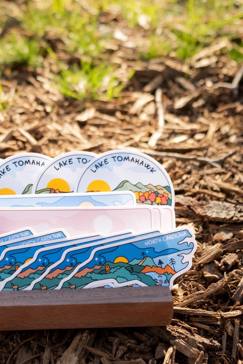 May include: A stack of stickers featuring landscape designs with text. The stickers have a variety of shapes and colors, including blue, pink, and white. Some stickers say "Lake Tomahawk" and "North Carolina".