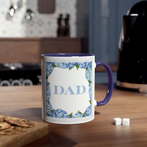 May include: White ceramic mug with a blue interior and handle. The mug features the word "DAD" in light blue, framed by a border of blue hydrangea flowers. The mug sits on a wooden surface.