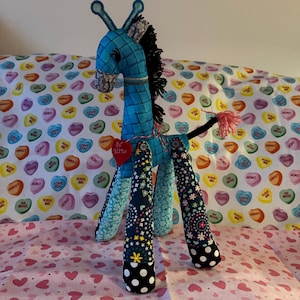 Op de afbeelding: Een handgemaakte knuffelgiraf met een blauw gewatteerd lichaam, zwarte manen en poten met patronen. De giraf heeft een rood hartvormig label en een roze kwaststaart. De achtergrond heeft een roze hartpatroon en een stof met snoephartontwerpen.