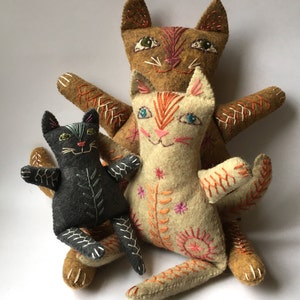 Könnte beinhalten: Drei handgefertigte Filzkatzenspielzeuge. Eine ist schwarz mit grünen Augen, eine ist braun mit orangen Augen und eine ist weiß mit orangen und blauen Augen. Alle drei Katzen haben gestickte Details.
