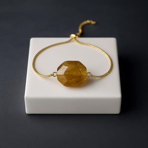 Peut inclure: Un bracelet doré avec une pierre précieuse facettée de couleur ambre au centre. Le bracelet réglable a un design de chaîne délicat. La pierre précieuse est de grande forme géométrique, et le bracelet est présenté sur une surface blanche sur fond sombre.