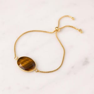 Peut inclure: Un bracelet doré avec une pierre d'oeil de tigre ovale et marron. Le bracelet est doté d'une chaîne délicate et d'un fermoir coulissant pour un ajustement facile. La pierre est sertie dans une lunette dorée.