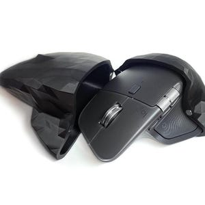 May include: A black computer mouse with a silver scroll wheel is housed in a black, geometric-patterned case. The mouse has a sleek, ergonomic design, and the case appears to be 3D-printed. The Logitech logo is visible on the mouse.