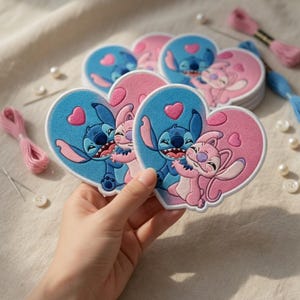 May include: Heart-shaped embroidered patches featuring the characters Stitch and Angel from Lilo & Stitch. The patches are blue and pink with a white border. The image also shows embroidery floss, needles, and buttons.