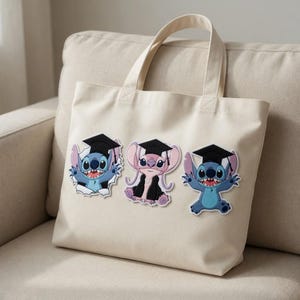 May include: Cream-colored tote bag featuring three cartoon characters wearing graduation caps. The characters are blue and pink, with black graduation caps and gowns. The bag has two handles and is sitting on a beige couch.