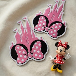 May include: Two pink castle-shaped patches with black and white polka dot bows. A Minnie Mouse figurine in a red dress with white polka dots, a red bow, and yellow shoes is also present. The patches and figurine are on a cream-colored surface.
