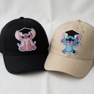 May include: Two baseball caps, one black and one tan, each featuring a cartoon character wearing a graduation cap. The black cap has a pink character, while the tan cap has a blue character. Both caps have a curved brim and a button on top.