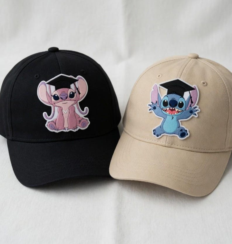 May include: Two baseball caps, one black and one tan, each featuring a cartoon character wearing a graduation cap. The black cap has a pink character, while the tan cap has a blue character. Both caps have a curved brim and a button on top.