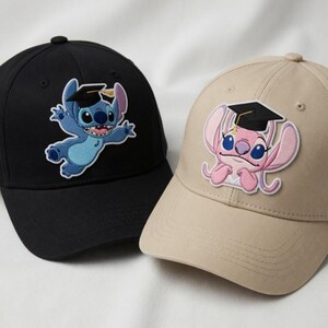 Puede incluir: Dos gorras de b&eacute;isbol con personajes de dibujos animados bordados. Una es negra con un personaje azul que lleva un birrete. La otra es beige con un personaje rosa que lleva un birrete. Ambas gorras tienen una visera curva.