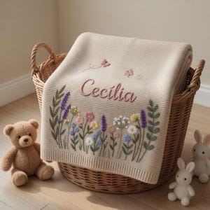 May include: A beige knitted baby blanket with the name "Cecilia" embroidered in pink, along with floral and butterfly designs. The blanket is displayed in a wicker basket, with a teddy bear and two toy bunnies nearby.