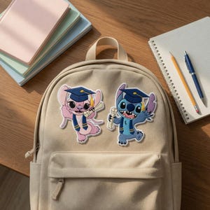 May include: A beige backpack with two embroidered patches of cartoon characters wearing graduation caps and gowns. One character is pink, the other is blue. A notebook, pen, and pencil are visible.