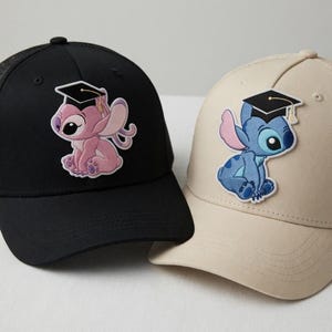 May include: Two baseball caps, one black and one tan, each featuring a cartoon character wearing a graduation cap. The black cap has a pink character, and the tan cap has a blue character. Both caps have a curved brim and a mesh back.