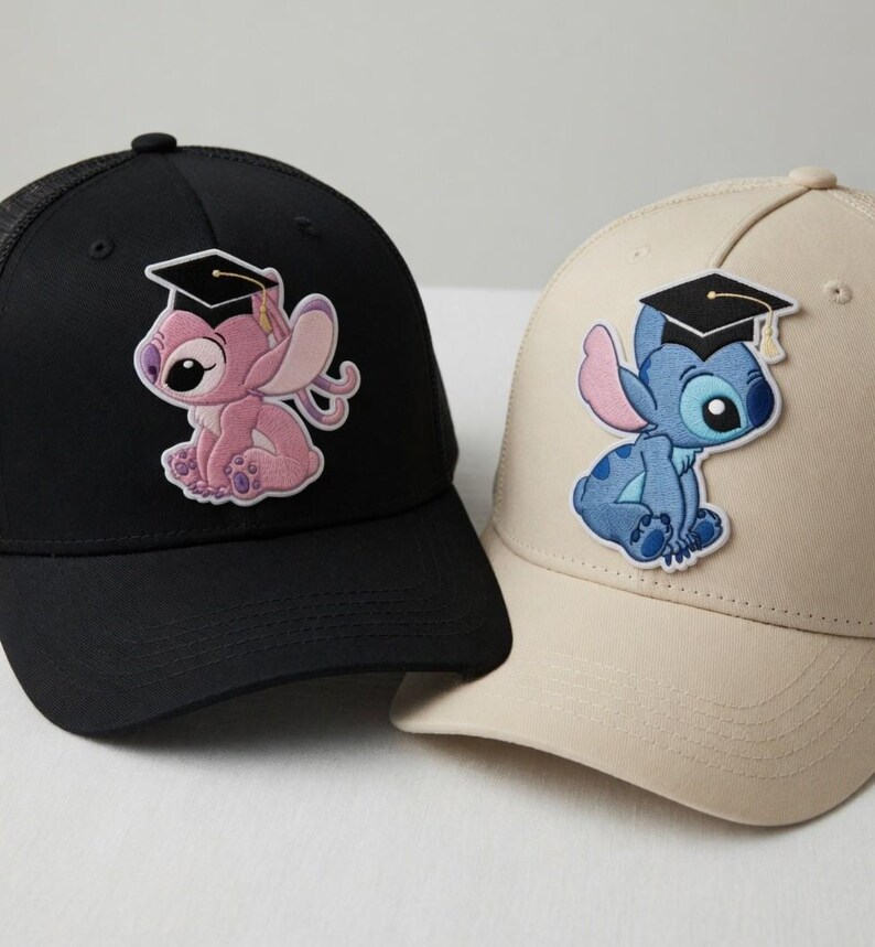 May include: Two baseball caps, one black and one tan, each featuring a cartoon character wearing a graduation cap. The black cap has a pink character, and the tan cap has a blue character. Both caps have a curved brim and a mesh back.