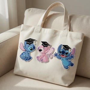 May include: Off-white canvas tote bag featuring three embroidered Stitch characters wearing graduation caps. The characters are in shades of blue and pink, with black caps and gold tassels. The bag has two handles.