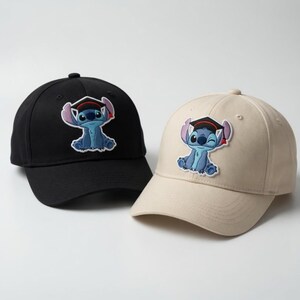 May include: Two baseball caps, one black and one beige, each featuring an embroidered patch of a blue cartoon character wearing a graduation cap. The caps have a curved brim and adjustable strap. The character is winking on the beige cap.