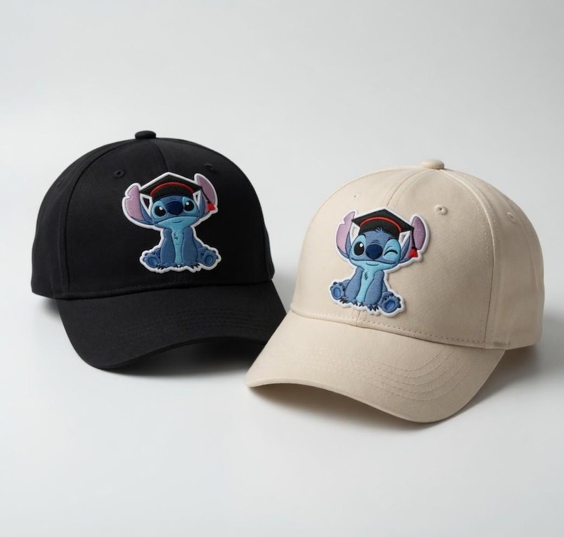 May include: Two baseball caps, one black and one beige, each featuring an embroidered patch of a blue cartoon character wearing a graduation cap. The caps have a curved brim and adjustable strap. The character is winking on the beige cap.