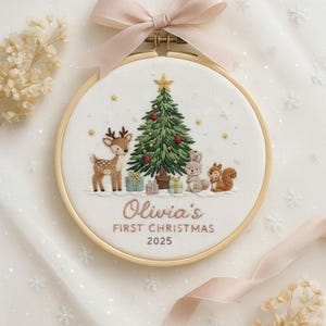 Puede incluir: Arte de aro navideño bordado con un árbol de Navidad, un ciervo, un conejo y una ardilla. El texto "Olivia's FIRST CHRISTMAS 2025" está bordado debajo. La obra está enmarcada en un aro de madera y tiene una cinta rosa.