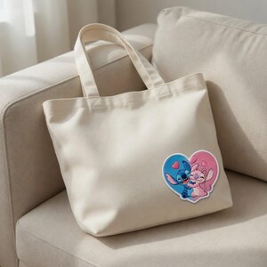 May include: A cream-colored tote bag with a heart-shaped patch featuring cartoon characters. The patch shows a blue character embracing a pink character within a blue and pink heart. The bag has two handles.