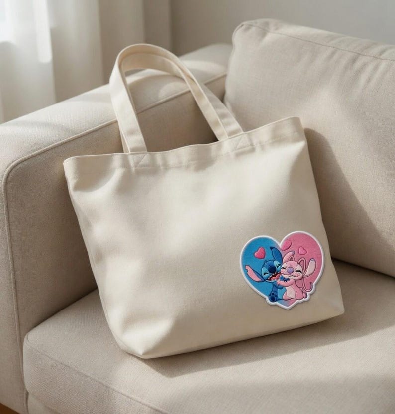 May include: A cream-colored tote bag with a heart-shaped patch featuring cartoon characters. The patch shows a blue character embracing a pink character within a blue and pink heart. The bag has two handles.