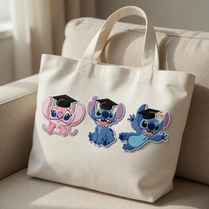 Puede incluir: Un bolso tote de color crema con tres personajes de dibujos animados bordados que llevan gorros de graduaci&oacute;n. Los personajes son en tonos de azul y rosa, con gorras negras y detalles amarillos. El bolso tiene dos asas.
