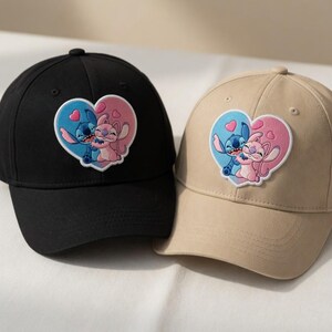 May include: Two baseball caps, one black and one tan, each featuring a heart-shaped patch with a cartoon couple embracing. The patch has a blue and pink color scheme, with the characters in the center. The caps have a curved brim and a button on top.
