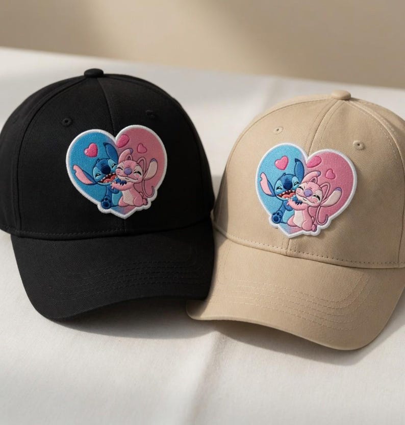 May include: Two baseball caps, one black and one tan, each featuring a heart-shaped patch with a cartoon couple embracing. The patch has a blue and pink color scheme, with the characters in the center. The caps have a curved brim and a button on top.