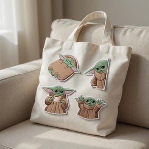 May include: A cream-colored tote bag with four embroidered patches of a green alien character in various poses. The patches are brown, green, and white. The bag has a simple design and a handle for carrying.