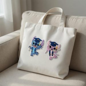 May include: A white tote bag featuring two cartoon characters, one blue and one pink, both wearing graduation caps and holding diplomas. The bag has two handles and is sitting on a beige sofa.