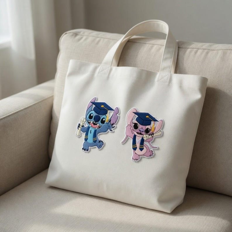 May include: A white tote bag featuring two cartoon characters, one blue and one pink, both wearing graduation caps and holding diplomas. The bag has two handles and is sitting on a beige sofa.