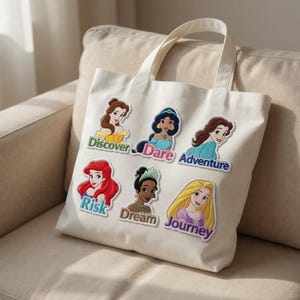 May include: Cream-colored tote bag featuring embroidered patches of animated female characters with text labels. The labels read "Discover," "Dare," "Adventure," "Risk," "Dream," and "Journey." The bag has two handles and is made of a canvas-like material.