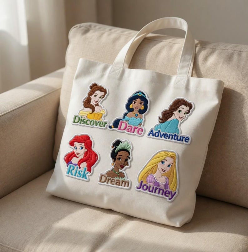 May include: Cream-colored tote bag featuring embroidered patches of animated female characters with text labels. The labels read "Discover," "Dare," "Adventure," "Risk," "Dream," and "Journey." The bag has two handles and is made of a canvas-like material.