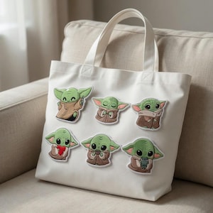 May include: White canvas tote bag with seven embroidered patches of a green alien character. The patches show the character in different poses, wearing a brown cloak and holding objects. The bag has two white handles and is on a beige sofa.
