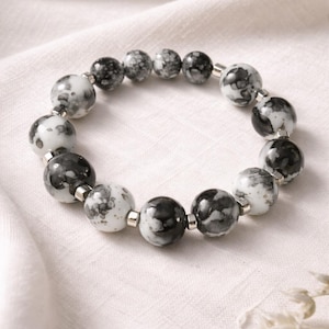 May include: A beaded bracelet featuring round, marbled beads in shades of black, white, and gray. The beads are strung together with silver-colored connectors, creating a stylish accessory. The bracelet is displayed on a white, textured surface.