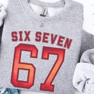 May include: Athletic gray sweatshirt with the text "SIX SEVEN" in a red and orange collegiate font, and the number "67" in a similar style. The sweatshirt is folded with the sleeves tucked in, and a silver necklace is draped across the front.