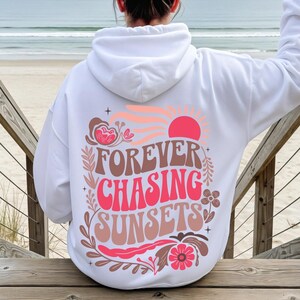 Pode incluir: Moletom branco com a frase "FOREVER CHASING SUNSETS" em um design retr&ocirc;. O design inclui um sol rosa, flores e um esquema de cores de p&ocirc;r do sol. O moletom &eacute; usado em um deck de madeira com vista para o oceano.