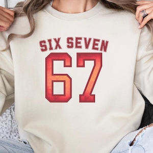 May include: A sand-colored sweatshirt with the text "SIX SEVEN" above the number "67" in a red and orange gradient. The word "SAND" is printed at the bottom of the image. The sweatshirt is a casual style.