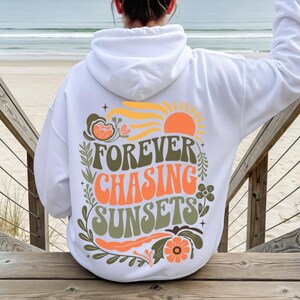 Pode incluir: Moletom branco com a frase "FOREVER CHASING SUNSETS" em laranja, verde e amarelo. O design inclui um sol, flores e folhas. A pessoa est&aacute; sentada em uma escada de madeira, com o mar ao fundo.