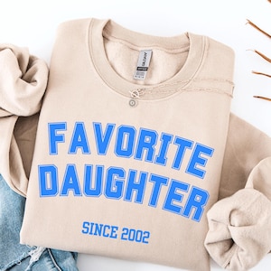 Pode incluir: Um moletom cor de areia com as palavras "FAVORITE DAUGHTER" em azul, com "SINCE 2002" abaixo. Um colar de prata com um pequeno pingente est&aacute; sobre a camisa. A palavra "SAND" est&aacute; na parte inferior.