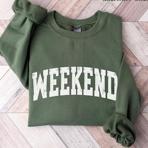 May include: A military green sweatshirt with the word "WEEKEND" printed in large, distressed white letters. The sweatshirt is folded, with the sleeves visible. The text "MILITARY GREEN" is in the top right corner.