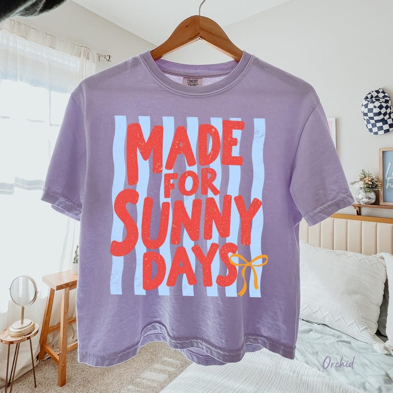 Peut inclure: Un crop top lavande avec le texte "MADE FOR SUNNY DAYS" en lettres rouges avec des rayures verticales bleu clair. Un motif de n&oelig;ud jaune est en bas. Le t-shirt est suspendu &agrave; un cintre en bois.