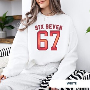 May include: White sweatshirt with the text "SIX SEVEN" above the number "67" in a red and orange gradient. The sweatshirt is a crew neck style and has long sleeves. The text is in a collegiate font.