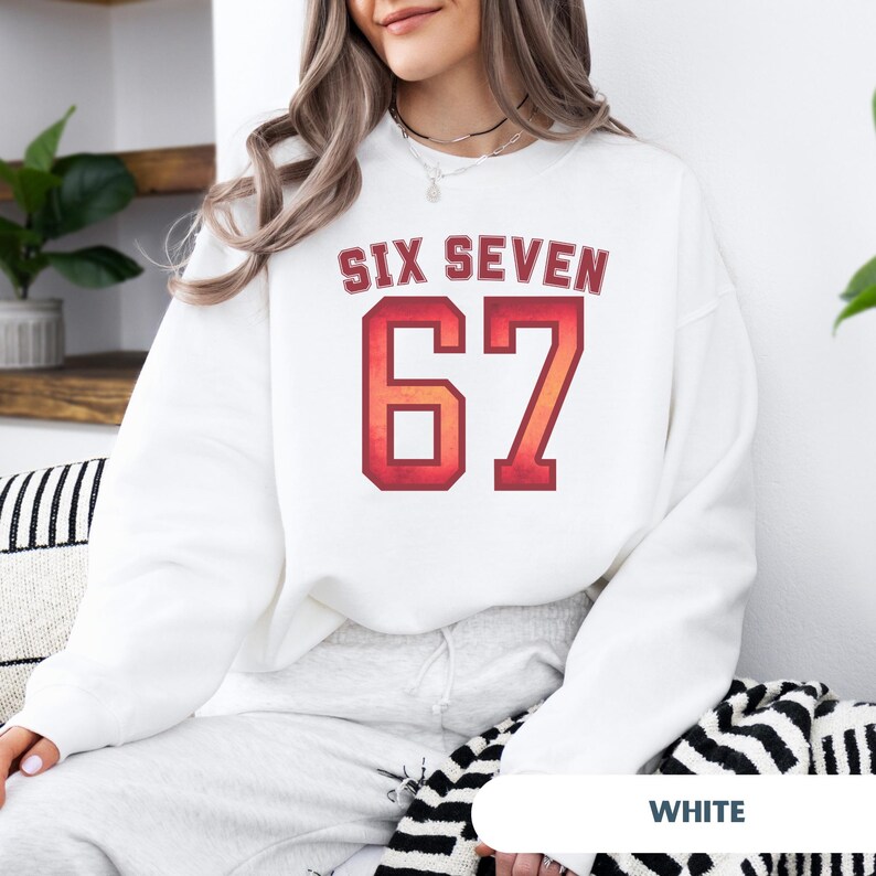 May include: White sweatshirt with the text "SIX SEVEN" above the number "67" in a red and orange gradient. The sweatshirt is a crew neck style and has long sleeves. The text is in a collegiate font.