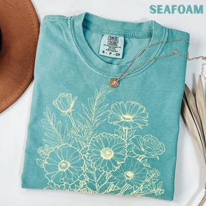 K&ouml;nnte beinhalten: Seegr&uuml;nes T-Shirt mit einem hellgelben Blumendesign. Das Shirt hat einen Rundhalsausschnitt und kurze &Auml;rmel. Eine goldene Halskette mit rundem Anh&auml;nger liegt auf dem Shirt. Der Text "SEAFOAM" steht oben.