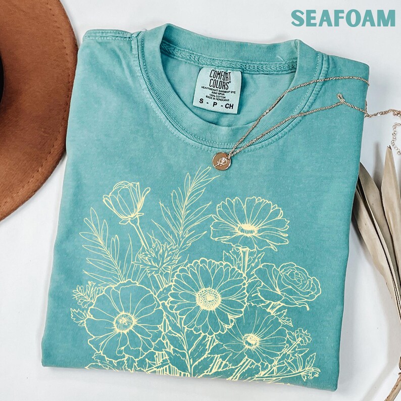 K&ouml;nnte beinhalten: Seegr&uuml;nes T-Shirt mit einem hellgelben Blumendesign. Das Shirt hat einen Rundhalsausschnitt und kurze &Auml;rmel. Eine goldene Halskette mit rundem Anh&auml;nger liegt auf dem Shirt. Der Text "SEAFOAM" steht oben.