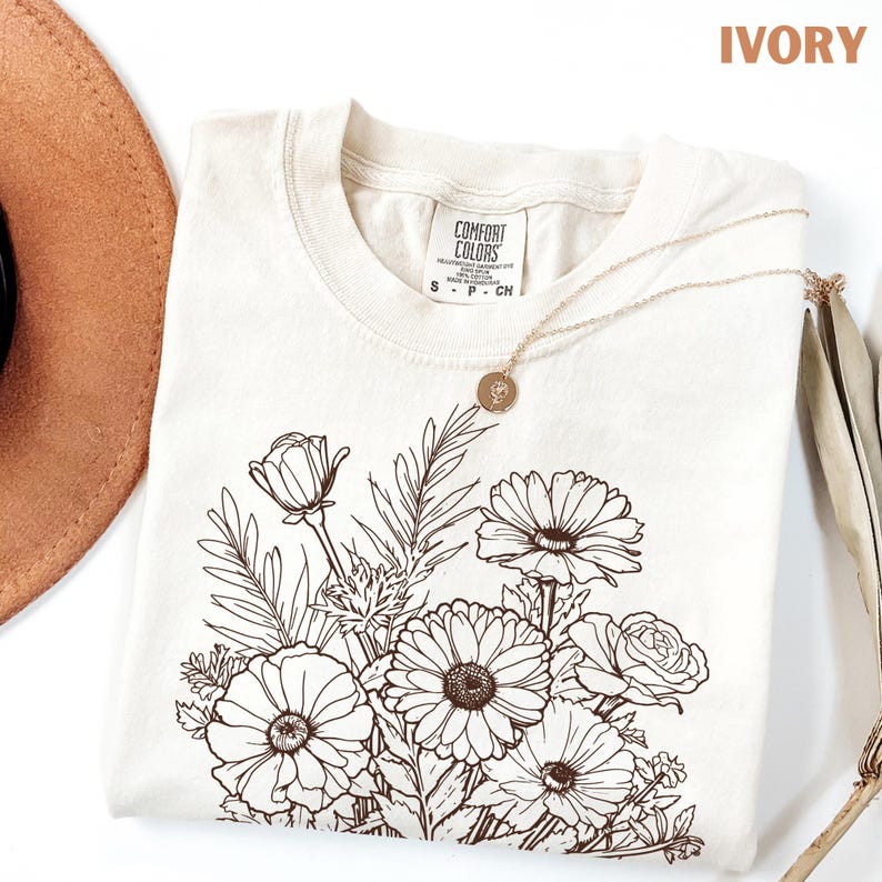K&ouml;nnte beinhalten: Elfenbeinfarbenes Comfort Colors T-Shirt mit braunem Blumendesign. Das T-Shirt hat einen Rundhalsausschnitt und kurze &Auml;rmel. Eine goldene Halskette mit Anh&auml;nger liegt auf dem Shirt. Ein brauner Hut ist teilweise sichtbar. Das Wort 'IVORY' ist sichtbar.