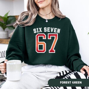 May include: Forest green sweatshirt with the text "SIX SEVEN" above the number "67" in a red and white design. The sweatshirt is a crew neck style. A white mug is held in the person's hand.
