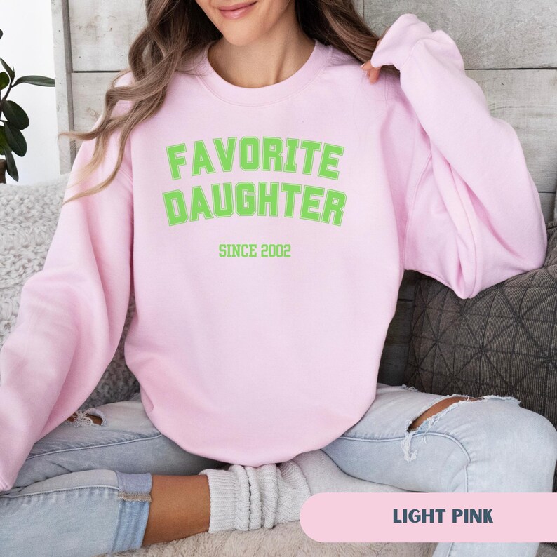 Pode incluir: Camisola rosa claro com a frase "FAVORITE DAUGHTER" em letras verdes brilhantes, com "SINCE 2002" abaixo. A camisola &eacute; de gola redonda. As palavras "LIGHT PINK" est&atilde;o na parte inferior da imagem.